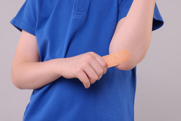 Little boy putting sticking plaster onto elbow against light grey background, closeup