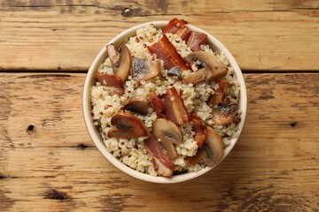 Tasty quinoa porridge with fried bacon and mushrooms in bowl on wooden table, top view