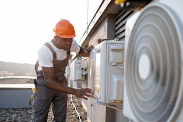 African american factory worker in orange hard hat standing and holding hands on air conditioner while checking attachment to wall outdoors. Concept of manual work and installation of equipment.