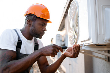 Focused african craftsman wearing orange hard hat holding screwdriver while screwing bolt into air conditioner. Serious specialist repairing broken device and performing work on rooftop. - obrazy, fototapety, plakaty