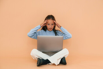 Worrying, sitting with laptop. Cute young girl is in the studio against background