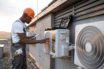 Qualified african american engineer looking at hanging air conditioner on rooftop of residential...