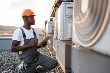 Focused man scrolling websites on digital tablet with useful information about air conditioners. African american craftsman in overalls and protective helmet looking at cooling system outdoors. - obrazy, fototapety, plakaty