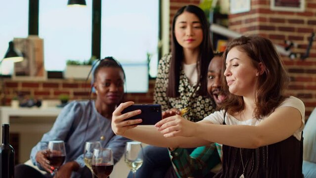 Asian, african american and caucasian people taking group pictures with mobile phone at apartment party. Friends in stylish home smiling for selfie photographs with wine and champagne glasses in hand
