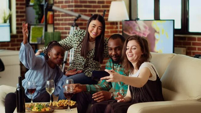 BIPOC friends taking group pictures with selfie camera smartphone at apartment party. Joyful mates in modern home posing for photo with wine and champagne glasses, making gestures