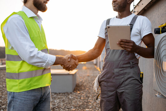 Friendly Young Multicultural Supervisors Shaking Hands On Rooftop Of Skyscraper. Ambitious Specialists Wearing Yellow Safety Vest And Gray Jumpsuit Holding Digital Tablet Outdoors.