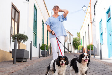 Smiling senior woman with hat walking in the street with her two cavalier king Charles spaniel...