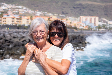 Fototapeta na wymiar Portrait of Happy Caucasian Senior Couple of Women Hugging in Vacation at Sea Standing Under the Sun Smiling