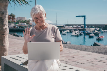 Video call concept. Active modern senior woman with glasses sitting at the harbor using laptop...