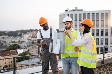 Friendly team of three project managers inspecting reduced copy of building that holding one of workers on rooftop with beautiful landscape on background. Concept of architecture and construction.