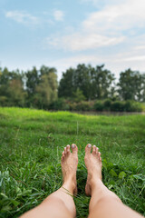 The image shows a pair of bare feet lying on a grassy field with a lake and a green park in the background. Toes point upward. The image is peaceful and calm.