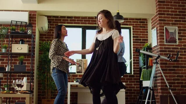 Happy friends dancing at apartment party, having fun together, rotating and jumping around. Mates doing energetic dance moves on upbeat songs in stylish living room, hip level shot