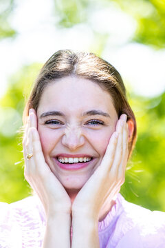 This image captures a young woman experiencing a moment of pure joy outdoors. She is holding her cheeks with her hands, and her expression is one of genuine delight and happiness. The woman is smile