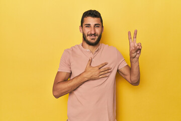 Young Hispanic man on yellow background taking an oath, putting hand on chest.