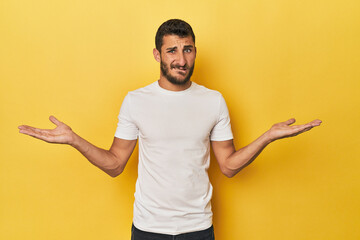 Young Hispanic man on yellow background confused and doubtful shrugging shoulders to hold a copy space. - obrazy, fototapety, plakaty