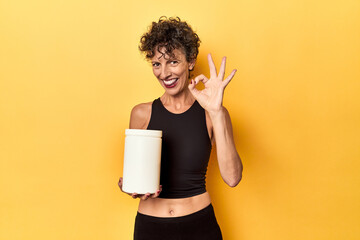 MIddle aged athlete woman holding protein supplement on yellow cheerful and confident showing ok...