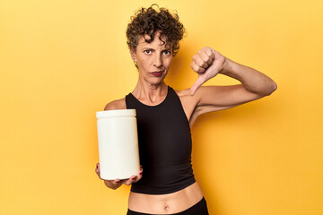 MIddle aged athlete woman holding protein supplement on yellow showing a dislike gesture, thumbs...