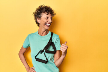 Sportswoman with resistance bands on yellow points with thumb finger away, laughing and carefree.