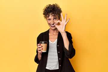 Businesswoman holding takeaway coffee on yellow cheerful and confident showing ok gesture.