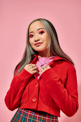 Valentines day, jolly asian young woman with heart eye makeup posing in red jacket on pink