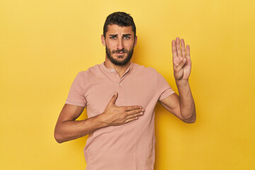 Young Hispanic man on yellow background taking an oath, putting hand on chest.