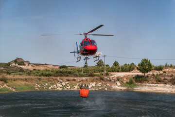 Helicóptero bombeiro a encher o balde que este trás suspenso por baixo num lago para o combate a...