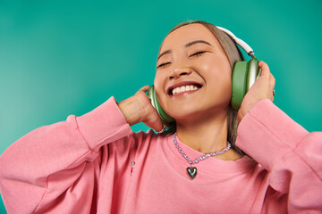 pleased young asian girl in wireless headphones listening music on turquoise background