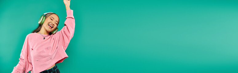positive young asian girl in wireless headphones gesturing and dancing on turquoise, banner