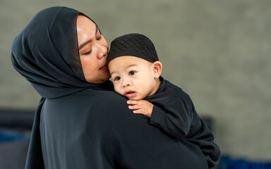 Portrait Adult muslim mother wearing traditional black dress and headscarf, carrying little baby boy, smiling with happiness in indoor home with copy space. Education, Family, Religious Concept.
