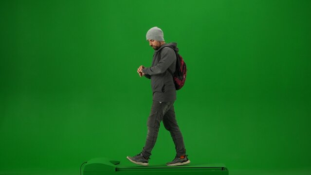 Portrait of person tourist isolated on chroma key green screen background. Adult man in knitted hat with backpack walking and looking at his watch.