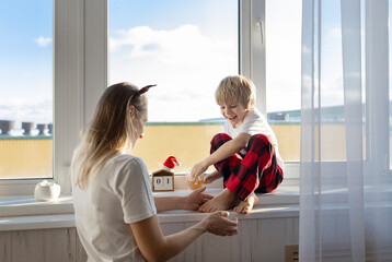 barefoot boy sits on the windowsill, with his mother tickling his feet. cozy family Christmas morning at home. cheerful start to a happy new year. concept of a friendly family, good mood - obrazy, fototapety, plakaty