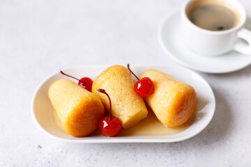 Neapolitan Rum baba (or baba au rum) on a white plate with a cocktail cherry and a cup of coffee. Small yeast cakes soaked in rum syrup on a gray background. Traditional Italian pastry. 