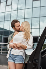 stylish handsome man embracing and kissing blonde girlfriend near modern car on urban street