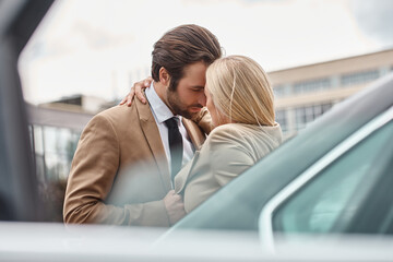 elegant business couple in formal wear embracing while standing near car on urban street, romance