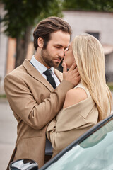 stylish businessman in formal wear embracing passionate blonde woman near car on city street