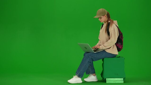 Portrait of traveler isolated on chroma key green screen background. Young girl sitting texting on laptop online, waiting for flight.
