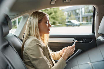 Fototapeta na wymiar side view of blonde businesswoman in formal wear messaging on mobile phone in luxury car on street