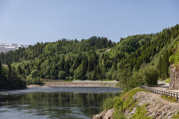 Fototapeta na wymiar A panoramic summer view of Snasavatnet, Norway's sixth-largest lake, framed by lush forests, a curving road, and a clear blue sky, capturing the essence of a vibrant, sunny day