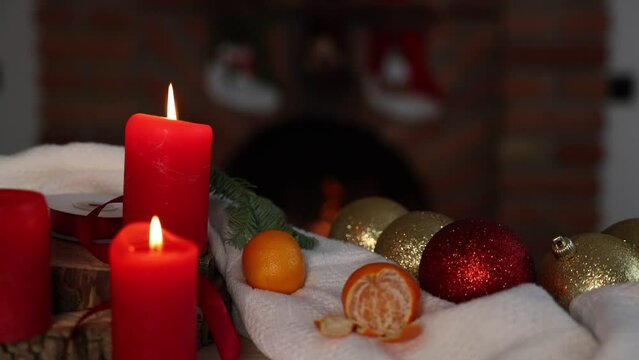 Candles flicker in the soft glow, accompanied by sparkling red and gold  balls scattered around them. A small pine tree branch complements the scene, and in the background a fireplace.