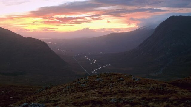 A winding river between mountain ranges illuminated by the rising sun. A82 road opposite Buachaille Etive Mor at dawn. Scotland. Panning forwards