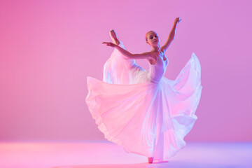 a young ballerina in pointe shoes dances in a long flowing white skirt on a pink background