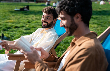 Happy ethnic gay couple reading books on deckchairs