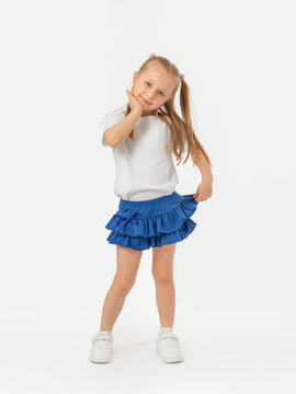 A cheerful 5-year-old girl posing on a white background with her hands folded under her chin in a white T-shirt, sneakers, blue skirt looks at the camera and smiles.