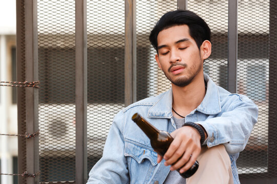 Alcoholic asian man drink beer from the bottle sitting on the floor after party. Sad heartbroken young man holding beer bottle in hand and get drunk till morning. - Powered by Adobe