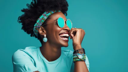 Poster Joyful woman with an afro hairstyle, laughing and wearing turquoise sunglasses against a vibrant turquoise background. © MP Studio