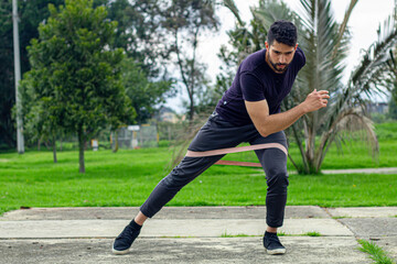 Obraz premium young man training with elastic bands, doing squat leg exercise outdoors in a park
