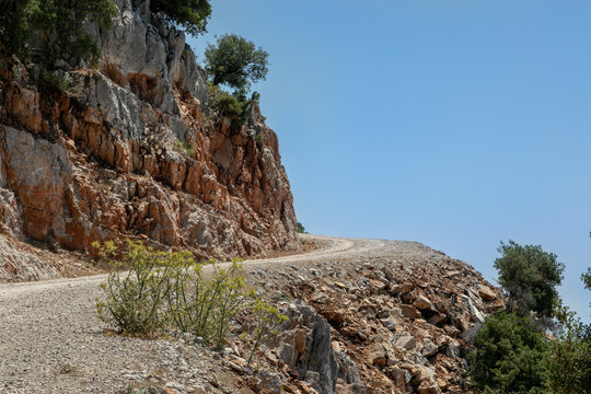 A sharp turn over the abyss. Dirt dusty road, steep rocky cliff, serpentine. The Mediterranean landscape is in the distance