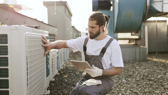 Focused master wearing gray overalls holding wireless tablet while inspecting air conditioner on plant. Caucasian qualified craftsman crouching and comparing information with device outdoors.