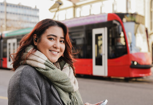 Happy young asian woman smiling on camera while using mobile phone during winter time in the city with tram in the background - Travel concept