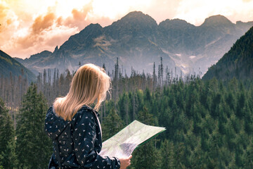A young girl on the background of forest, mountains and red sky looking at a map - obrazy, fototapety, plakaty